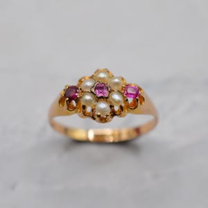 Pode incluir: Um anel de ouro com um design central em cacho. A cravação inclui quatro pequenas pérolas brancas dispostas em torno de uma pedra preciosa rosa central de corte quadrado, ladeada por duas pedras preciosas rosa adicionais nas laterais.