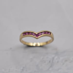 Puede incluir: Un anillo dorado con un diseño en forma de V. El anillo está engastado con una fila de pequeñas piedras preciosas de color rosa vibrante. La banda tiene un acabado liso y pulido, y las piedras preciosas están espaciadas uniformemente.