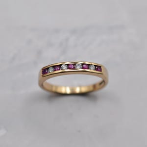 May include: A gold band ring with alternating red gemstones and clear gemstones. The ring is set with small, round-cut stones. The ring is on a light gray background.