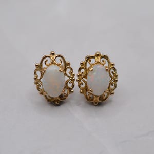 May include: A pair of gold-toned stud earrings. Each earring features an oval, iridescent opal stone set within a decorative, filigree frame. The earrings are displayed on a light-colored surface.