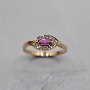 Può includere: Un anello d'oro con una gemma rosa a forma di marquise, circondata da piccole gemme trasparenti. La fascia dell'anello ha una curva delicata, creando un design elegante e classico. L'anello è posto su uno sfondo neutro.