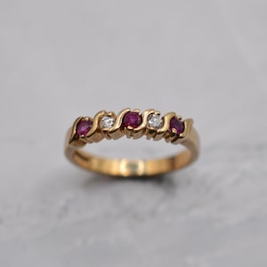 May include: A gold ring featuring a series of alternating red gemstones and clear stones. The ring has a curved design, with the gemstones set in a wave-like pattern. The ring is set against a neutral background.