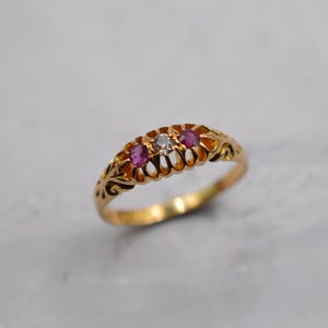 May include: A gold ring with three gemstones. The ring features two red gemstones and one clear gemstone in the center. The band has an ornate, filigree design. The ring is set against a neutral background.