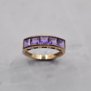 Puede incluir: Un anillo de oro con cinco piedras preciosas moradas de corte cuadrado. El anillo tiene un diseño sencillo y elegante. Las piedras están dispuestas en una fila, creando un aspecto clásico y atemporal.