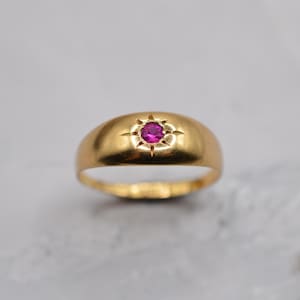 May include: A gold ring featuring a rounded band and a central, starburst-set, round, magenta gemstone. The ring has a polished finish, reflecting light. The gemstone is the focal point, set within a detailed starburst design.