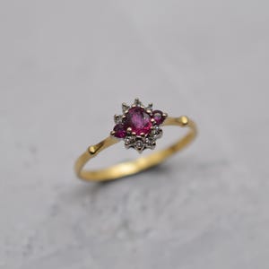 Op de afbeelding: Een gouden ring met een cluster van edelstenen. De centrale ovale robijn is omgeven door kleinere robijnen en heldere accentstenen. De band heeft een delicaat, getextureerd ontwerp.