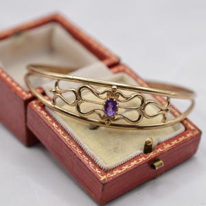 May include: A gold-coloured bangle bracelet featuring an oval, purple gemstone. The bracelet has an openwork design with decorative swirls. The bracelet is displayed in a red jewellery box with a cream-coloured interior.