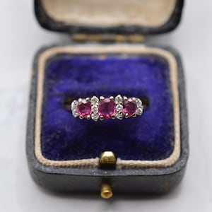 May include: A silver-toned ring featuring three oval-cut pinkish-red gemstones arranged horizontally, interspersed with small clear round-cut stones. The ring is displayed inside a dark-colored jewelry box with a deep blue velvet interior lining.