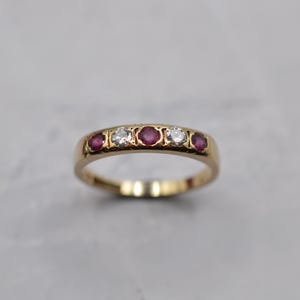 May include: A gold ring with five gemstones. It has alternating round, red rubies and clear, round diamonds. The ring is set in a gold band.
