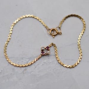 Könnte beinhalten: Ein goldenes Kettenarmband mit einem quadratischen Mittelstück, das mit zwei runden klaren Steinen und zwei runden roten Steinen besetzt ist. Das Armband hat ein sich wiederholendes S-Glied-Muster und einen runden Federringverschluss.