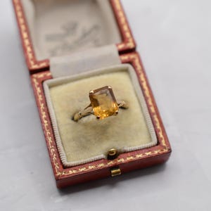 May include: A gold ring with a rectangular, amber-colored gemstone is displayed inside a red jewelry box with a cream-colored interior. The ring's band has a delicate, ornate design. The box is open, revealing the ring's setting.