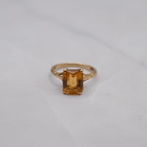 May include: A gold ring with a rectangular, amber-colored gemstone. The ring's band is a simple, classic design, and the gemstone is set in a four-prong setting. The ring is likely a piece of jewelry, suitable for everyday wear or special occasions.