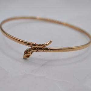Może przedstawiać: Bransoletka typu bangle z metalu w kolorze różowego złota z motywem węża. Głowa węża posiada teksturowane łuski oraz dwa małe, jaskrawopomarańczowe okrągłe kamienie jako oczy. Ciało węża owija się wokół, tworząc zapięcie bransoletki.