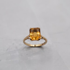May include: A gold ring featuring a large, rectangular, amber-colored gemstone. The ring has a delicate, vintage design with a slender band and decorative accents on the sides of the gemstone setting. The gemstone is the focal point.