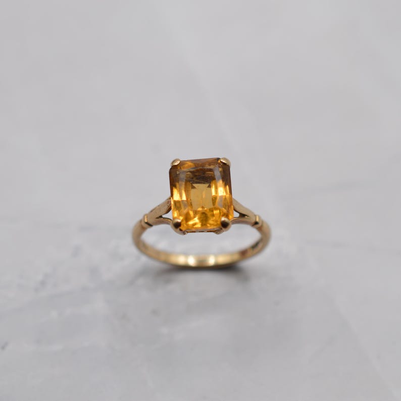 May include: A gold ring featuring a large, rectangular, amber-colored gemstone. The ring has a delicate, vintage design with a slender band and decorative accents on the sides of the gemstone setting. The gemstone is the focal point.