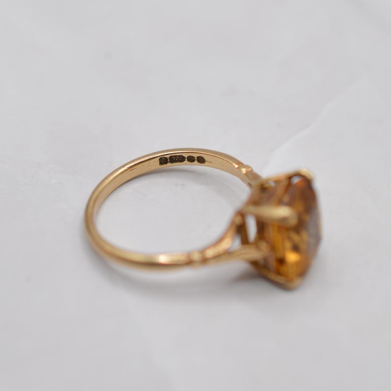 May include: A gold ring with an amber-colored gemstone. The ring has a detailed band and a large, faceted stone. The ring is set against a white background, highlighting its color and design. The ring is likely a vintage or antique piece.