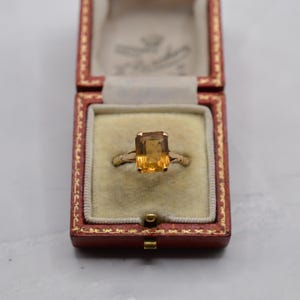 May include: A gold ring with a rectangular, amber-colored gemstone, set in a vintage red jewelry box. The ring's band is a delicate gold, and the gemstone is the focal point.