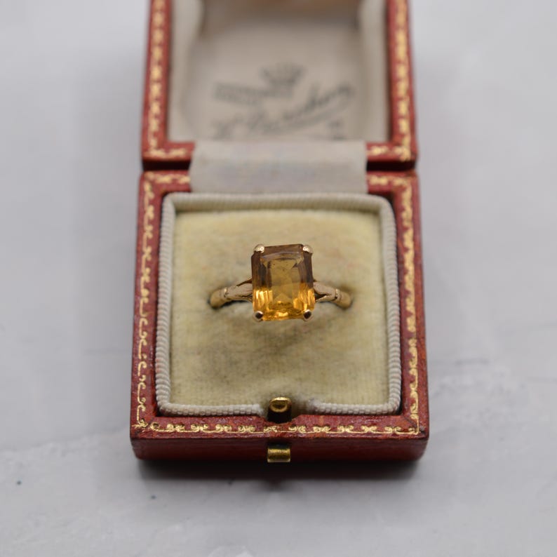 May include: A gold ring with a rectangular, amber-colored gemstone, set in a vintage red jewelry box. The ring's band is a delicate gold, and the gemstone is the focal point.