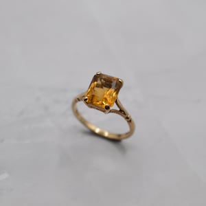 May include: A gold ring featuring a rectangular, amber-colored gemstone. The ring's band has an ornate design with delicate detailing. The gemstone is securely set within the ring's setting, showcasing its warm, golden hue. This piece of jewelry is a classic and elegant accessory.