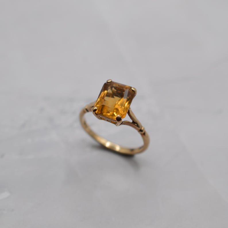 May include: A gold ring featuring a rectangular, amber-colored gemstone. The ring's band has an ornate design with delicate detailing. The gemstone is securely set within the ring's setting, showcasing its warm, golden hue. This piece of jewelry is a classic and elegant accessory.