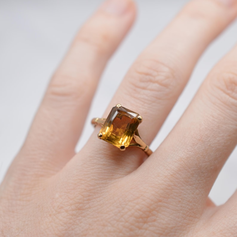 May include: A gold ring featuring a large, rectangular, amber-colored gemstone. The ring's band has an ornate design, and the gemstone is set in a classic, elegant style. The ring is shown on a person's finger.