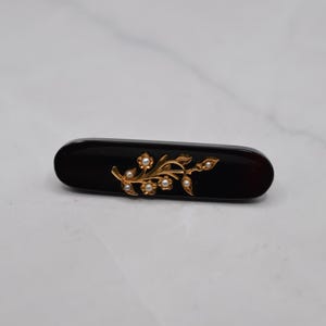 May include: A dark brown, oval-shaped hair barrette with a gold-colored floral design. The floral design features small, white pearl-like accents. The barrette is approximately 3 inches long.
