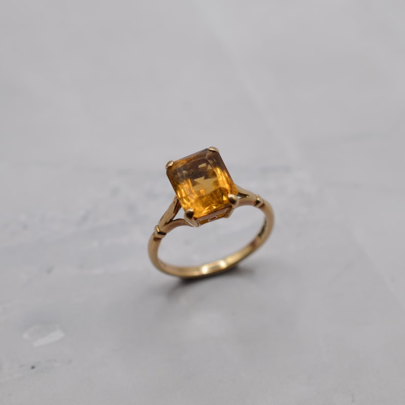 May include: A gold ring featuring a rectangular, amber-colored gemstone. The ring has a delicate, ornate design with a slender band. The gemstone is the focal point, catching the light and showcasing its warm, golden hue. A classic piece of jewelry.