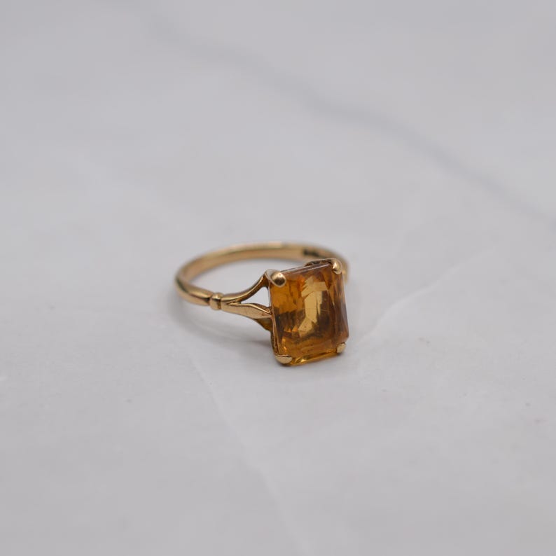 May include: A gold ring with a rectangular, amber-colored gemstone. The ring's band has a delicate design with a split shank. The gemstone is set in a classic, four-prong setting. The ring is displayed on a white surface.