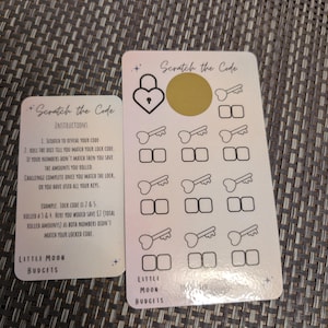 May include: Two white cards with the text "Scratch the Code" and instructions. One card has a gold scratch-off circle and key and lock graphics. The other card has instructions and the Little Moon Budgets logo.