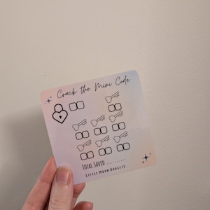 May include: A small, square card with a pastel gradient background is held in a hand. The card features the text "Crack the Mini Code" and illustrations of locks, keys, and boxes. The words "TOTAL SAVED" and "LITTLE MOON BUDGETS" are also present.