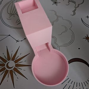 May include: A light pink 3D-printed object with a rectangular top section and a circular base. The object has a curved connecting piece between the two sections. The object is on a grey surface with celestial designs.