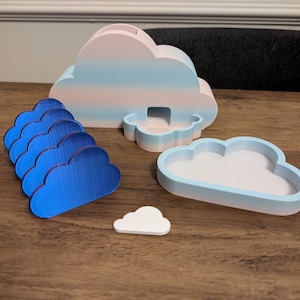 May include: A set of cloud-themed desk accessories. Features a striped pink and blue pen holder, a small tray, a stack of blue cloud cutouts, and a small white cloud. The items are made of wood or plastic and are designed to look like clouds.