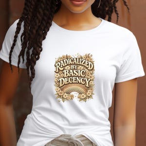 May include: White t-shirt featuring the text "RADICALIZED BY BASIC DECENCY" in a vintage-style design. The graphic includes a rainbow and floral elements in shades of brown, beige, and green.