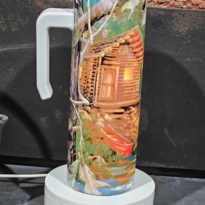May include: A white and green camouflage patterned stainless steel tumbler with a white handle. The tumbler features a cabin in the woods with a red canoe in the water.
