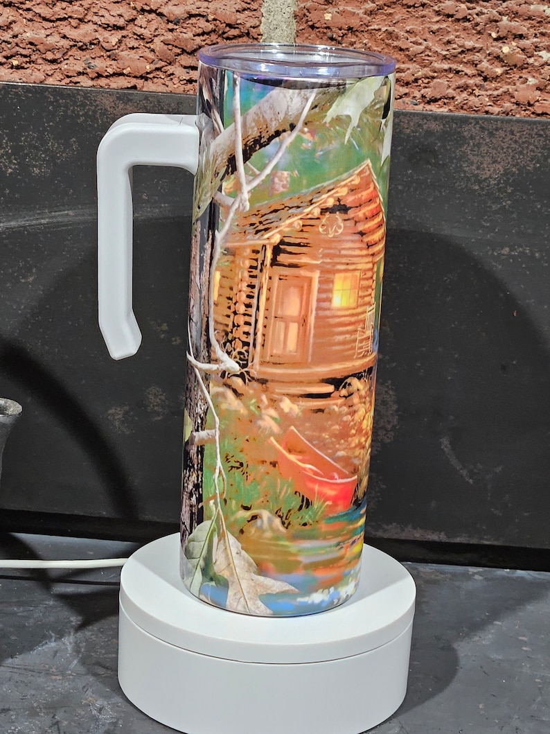 May include: A white and green camouflage patterned stainless steel tumbler with a white handle. The tumbler features a cabin in the woods with a red canoe in the water.