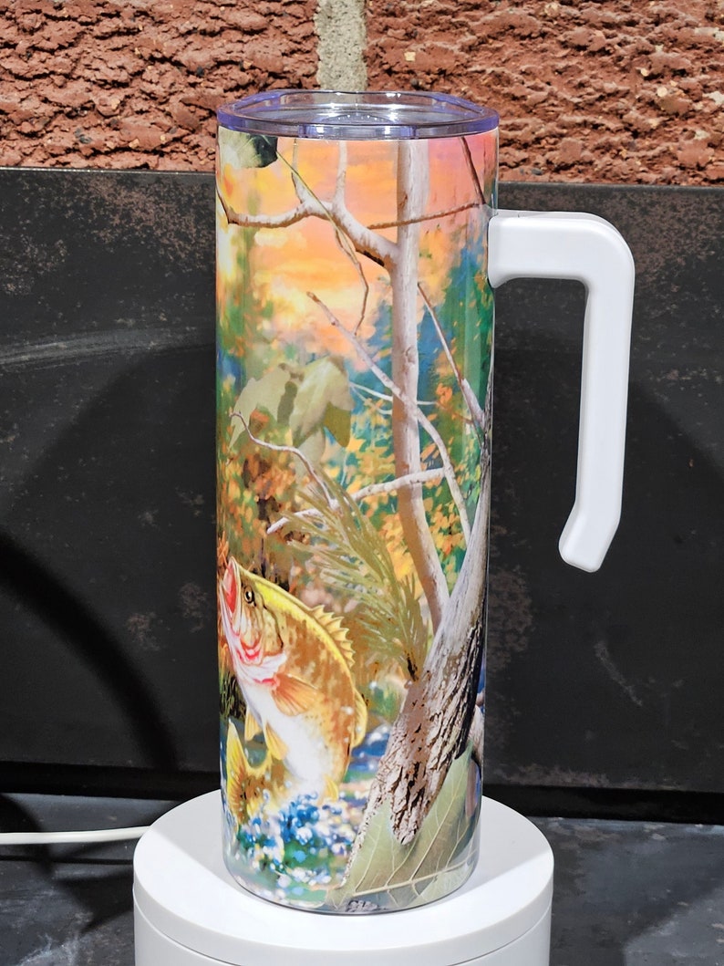 May include: A white stainless steel travel mug with a white handle. The mug has a colorful print of a fish jumping out of the water in a forest setting.
