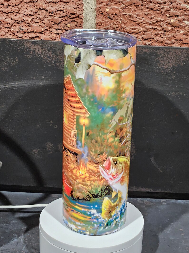 May include: A stainless steel tumbler with a colorful design featuring a cabin in the woods, a campfire, and a largemouth bass jumping out of the water. The design is reminiscent of a fishing scene.