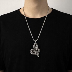 May include: Silver-toned dragon pendant necklace. The detailed dragon design is suspended from a matching chain. The necklace is worn against a black shirt.