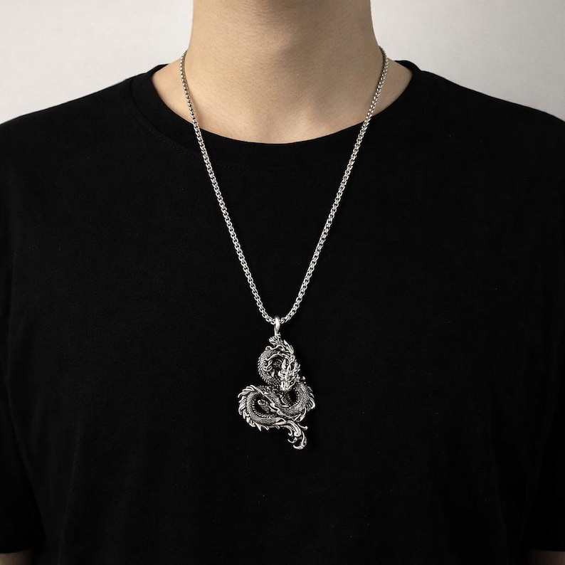 May include: Silver-toned dragon pendant necklace. The detailed dragon design is suspended from a matching chain. The necklace is worn against a black shirt.
