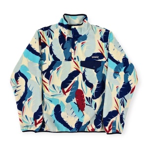 May include: A cream-colored fleece pullover with a vibrant leaf pattern in shades of blue, teal, and red. Features a navy blue pocket and collar, and a button-up closure. The Patagonia logo is visible on the pocket.