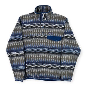 May include: A patterned fleece pullover in shades of blue, gray, and white. The geometric design covers the entire garment, including the high collar and a small chest pocket. The pullover has a buttoned placket and a small brand label.