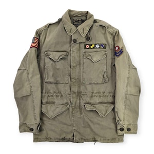 Puede incluir: Chaqueta de estilo militar verde oliva con múltiples bolsillos y cierres de botón. La chaqueta presenta un parche de la bandera estadounidense en la manga izquierda y varios parches bordados en el pecho. Cuello con solapas y cintura ajustable.