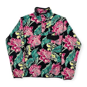 May include: A black fleece pullover with a vibrant floral pattern featuring pink, green, and yellow flowers and leaves. The pullover has a stand-up collar, a snap closure, and a small black pocket on the chest. The Patagonia logo is visible on the chest.