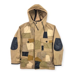 May include: A tan hooded jacket with a patchwork design. The jacket features various patches in shades of tan, beige, black, and olive green. Black leather patches are on the elbows. The jacket has a drawstring hood and multiple pockets.