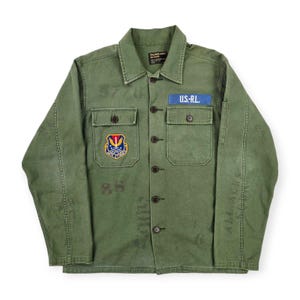 Puede incluir: Chaqueta verde oliva de estilo militar con cierre de botones, dos bolsillos en el pecho y cuello. Cuenta con un parche azul que dice "U.S.-R.L." y un parche colorido con un escudo. La chaqueta tiene marcas descoloridas y está hecha de tela duradera.