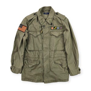 Può includere: Giacca verde oliva in stile militare con tasche multiple e toppe. Presenta una toppa con la bandiera americana sulla manica sinistra e altre insegne. La giacca ha una chiusura con bottoni e un colletto.