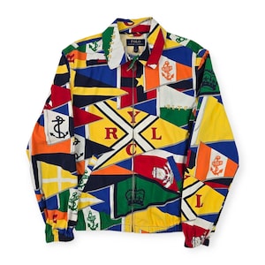May include: A zip-up jacket featuring a vibrant all-over print of nautical signal flags in red, yellow, blue, green, and white. The design includes anchor motifs, crowns, and the letters R, L, Y, and C. It has a collar, long sleeves with elastic cuffs, and a Polo Ralph Lauren label at the neck.