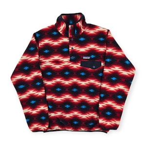 May include: A patterned fleece pullover in red, black, cream, and blue. The pullover features a high collar, a buttoned placket, and a chest pocket. The design includes geometric shapes and the brand's logo.