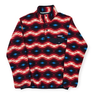 May include: A red, black, and cream-colored fleece pullover with a geometric pattern. The pullover has a navy blue pocket and collar, and a small brand patch. It features a button-up closure and a high collar.