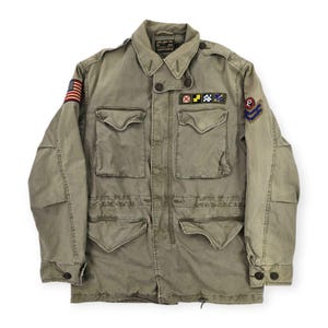 May include: An olive green field jacket featuring a button-up front, four large front pockets, and a collar. The right sleeve displays an American flag patch, while the left chest has a row of signal flag patches and the left sleeve has a chevron patch.